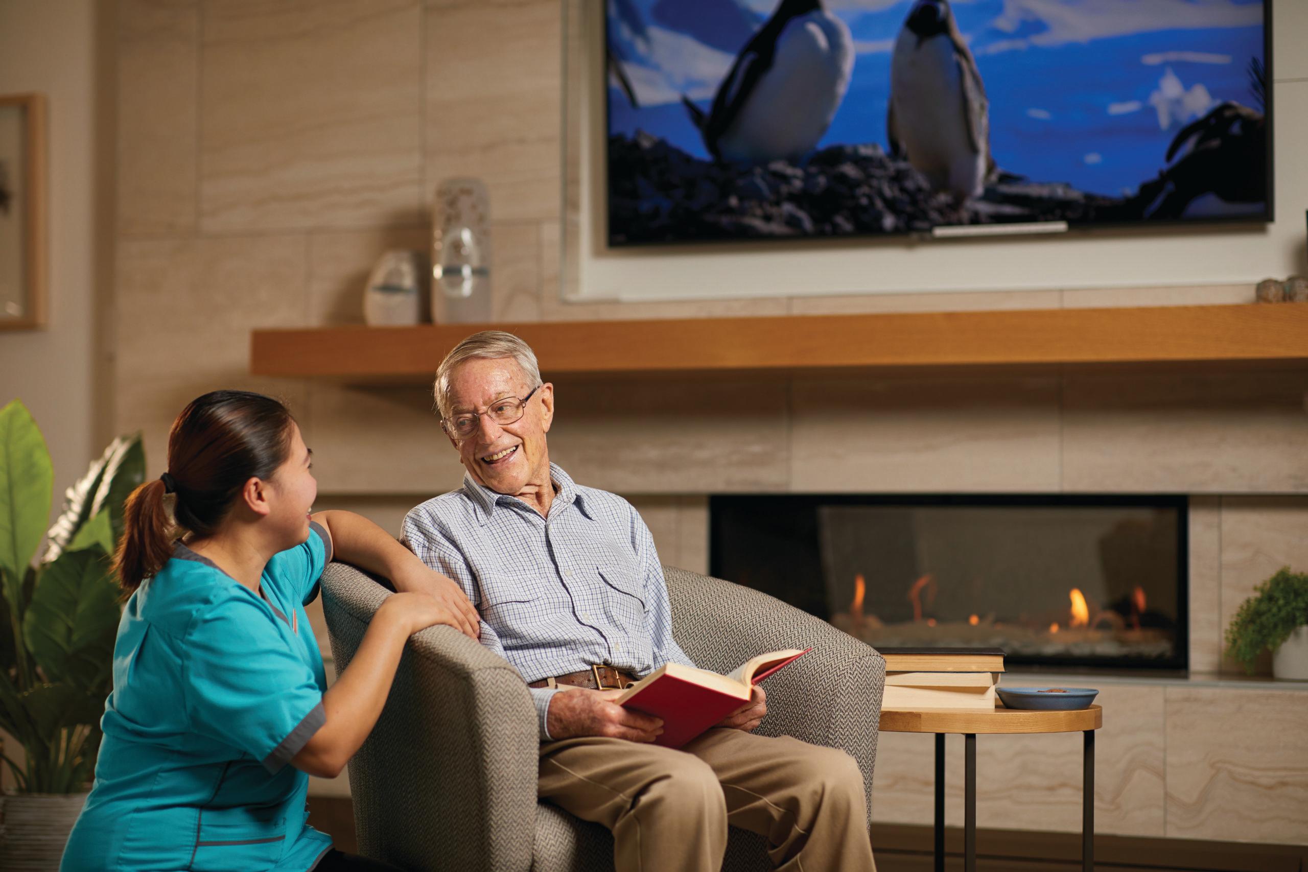 Aged care resident with Healthcare Assistant by fireplace reading book